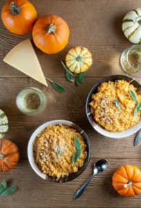 2 bowls of pumpkin risotto on wood with small pumpkins, glasses of white wine, and parmesan cheese