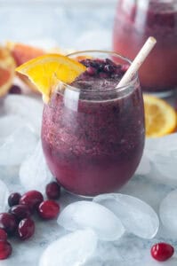 two pomegranate cranberry smoothies on white marble with ice cubes, orange slices, and frozen cranberries