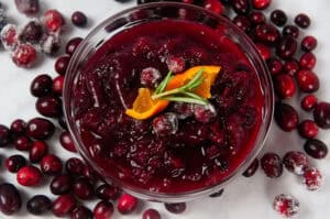 A clear bowl of Instant Pot Cranberry Sauce surrounded by cranberries on white. It is a bright, citrusy holiday side dish.