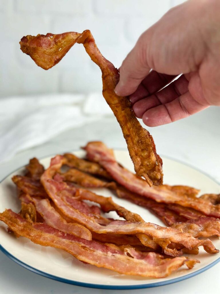 A hand picking up a crispy piece of bacon off a plate