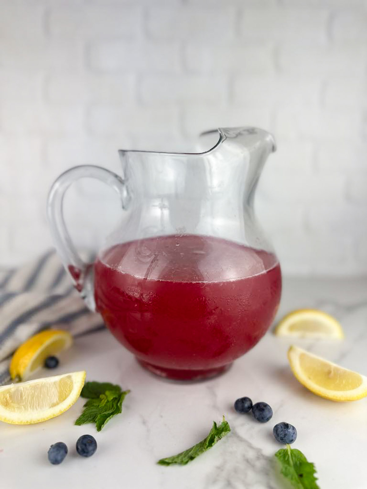 A pitcher of blueberry lemonade with some lemon wedges, blueberries, and mint leaves