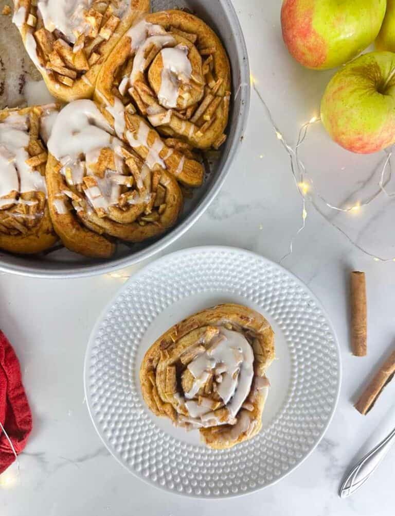 A plate with an Apple Pie Cinnamon Roll in front of a cake pan full of Apple Pie Cinnamon Rolls