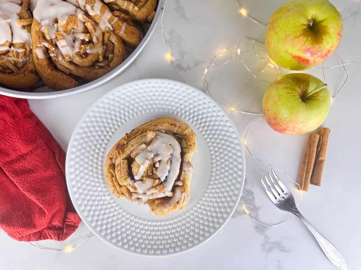 A plate with an Apple Pie Cinnamon Roll in front of a cake pan full of Apple Pie Cinnamon Rolls