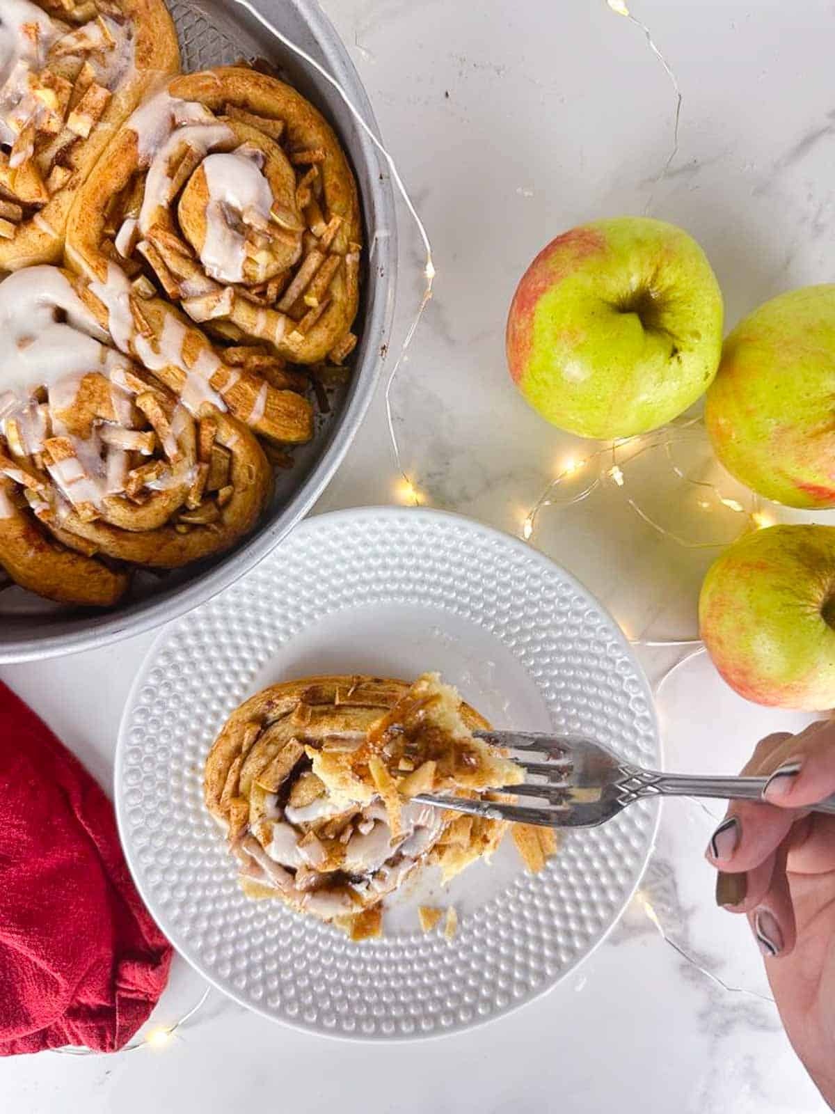 A plate with an Apple Pie Cinnamon Roll in front of a cake pan full of Apple Pie Cinnamon Rolls