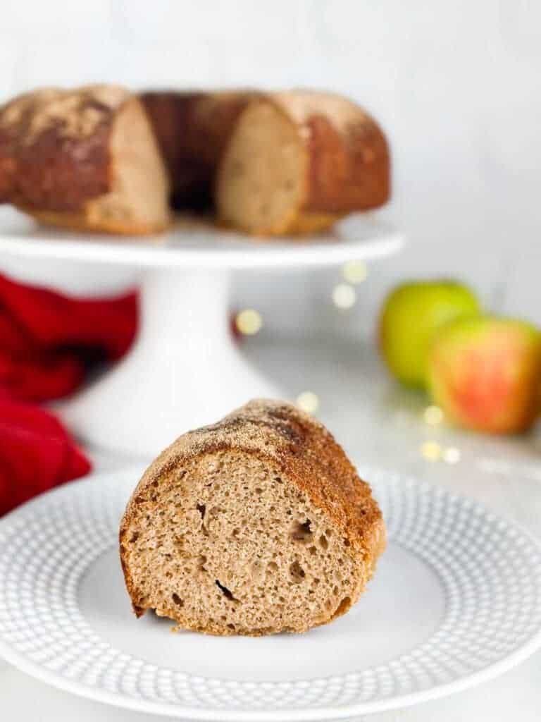 A slice of apple cider cake on a plate in front of an apple cider bundt cake