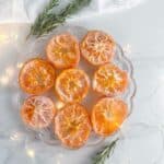 Plate of candied oranges dipped in sugar on a plate with rosemary twigs and twinkle lights surrounding it.