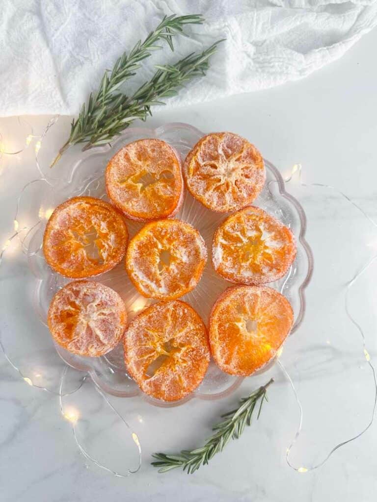 Plate of candied oranges dipped in sugar on a plate with rosemary twigs and twinkle lights surrounding it.