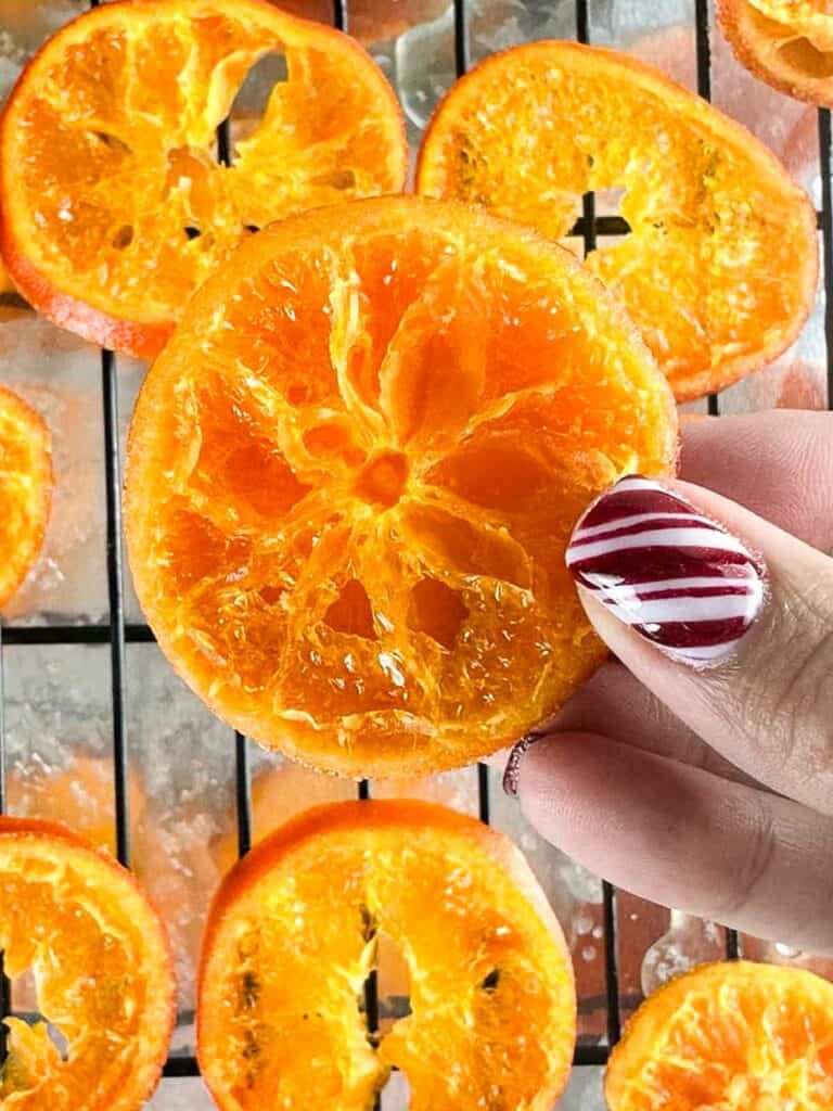 Hand holding baked candied orange slice over baked orange slices on cooling rack over a foil lined sheet.