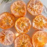 Pictured candied orange slices on a clear plate with twinkle lights and rosemary twigs on the sides; text reads candied orange slices.