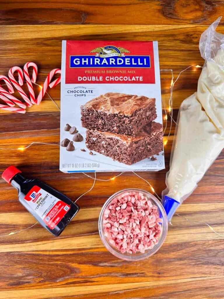 Ingredients for candy cane brownies spread out on counter: box of Ghirardelli double chocolate brownies, peppermint extract, peppermint frosting in a piping bag, and crushed candy canes.