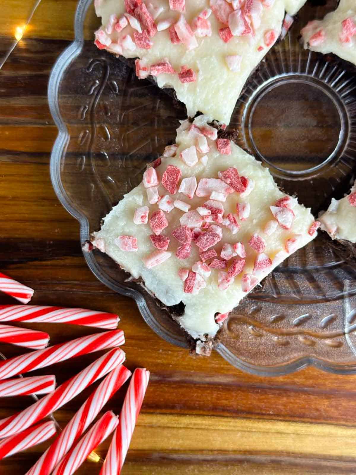 Plate of four candy cane brownies with some candy canes in the foregrounds and white twinkle lights on top of a butcher block counter.