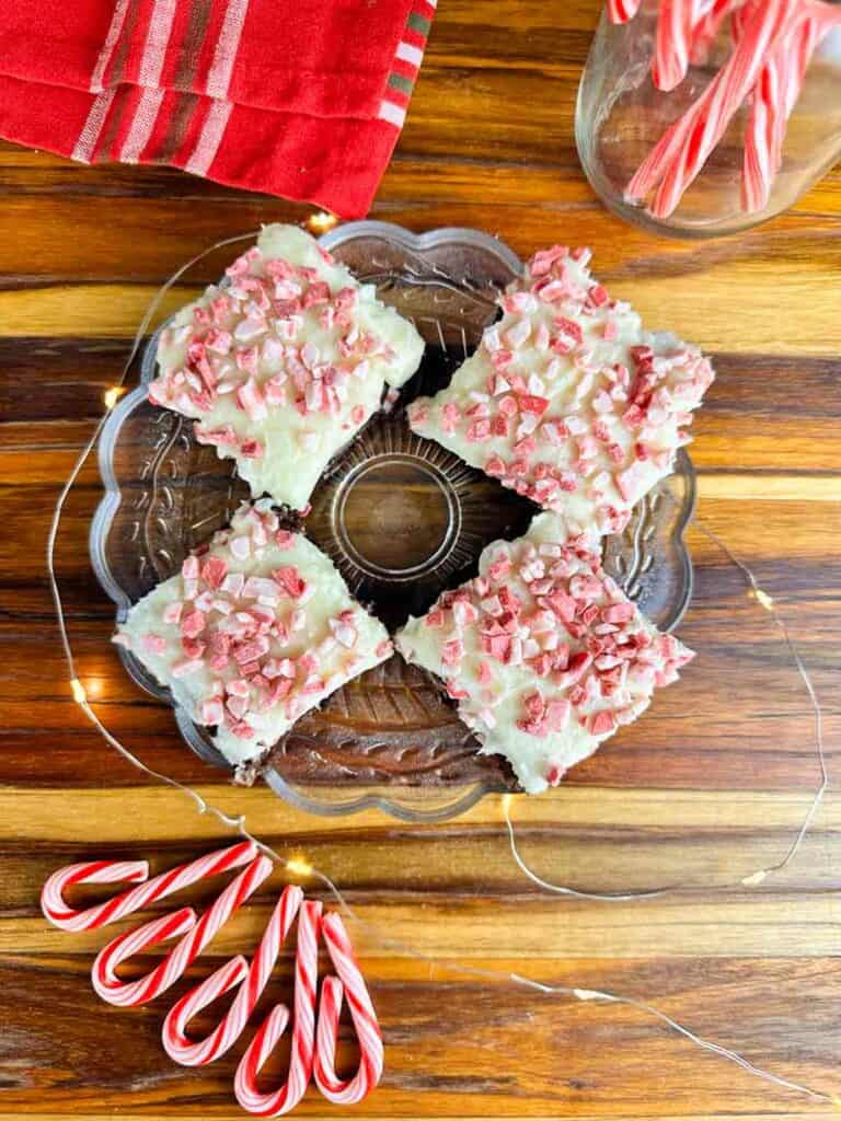 Plate of four candy cane brownies with some candy canes in the foregrounds and white twinkle lights on top of a butcher block counter.