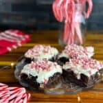 Plate of four candy cane brownies with a jar of candy canes in the background, some candy canes in the foreground, and a red tea towel all on top of a butcher block counter.
