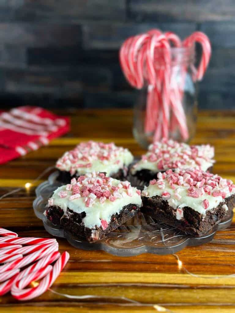 Plate of four candy cane brownies with a jar of candy canes in the background, some candy canes in the foreground, and a red tea towel all on top of a butcher block counter.