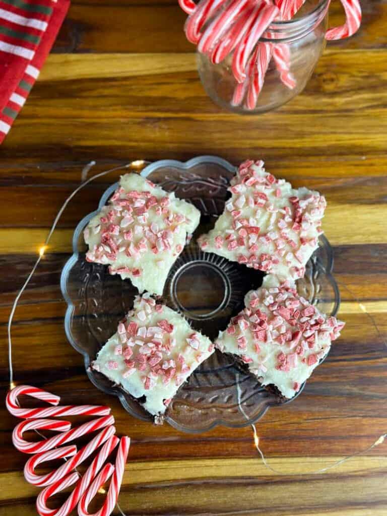 Plate of four candy cane brownies with a jar of candy canes in the background, some candy canes in the foreground, and a red tea towel all on top of a butcher block counter.