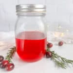 Mason jar with lid with cranberry simple syrup. Cranberries, rosemary sprigs, a tea towel, and twinkle lights are all in the background.