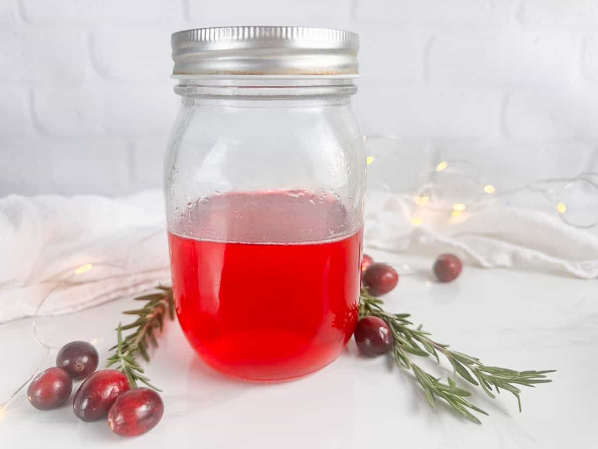 Mason jar with lid with cranberry simple syrup. Cranberries, rosemary sprigs, a tea towel, and twinkle lights are all in the background.