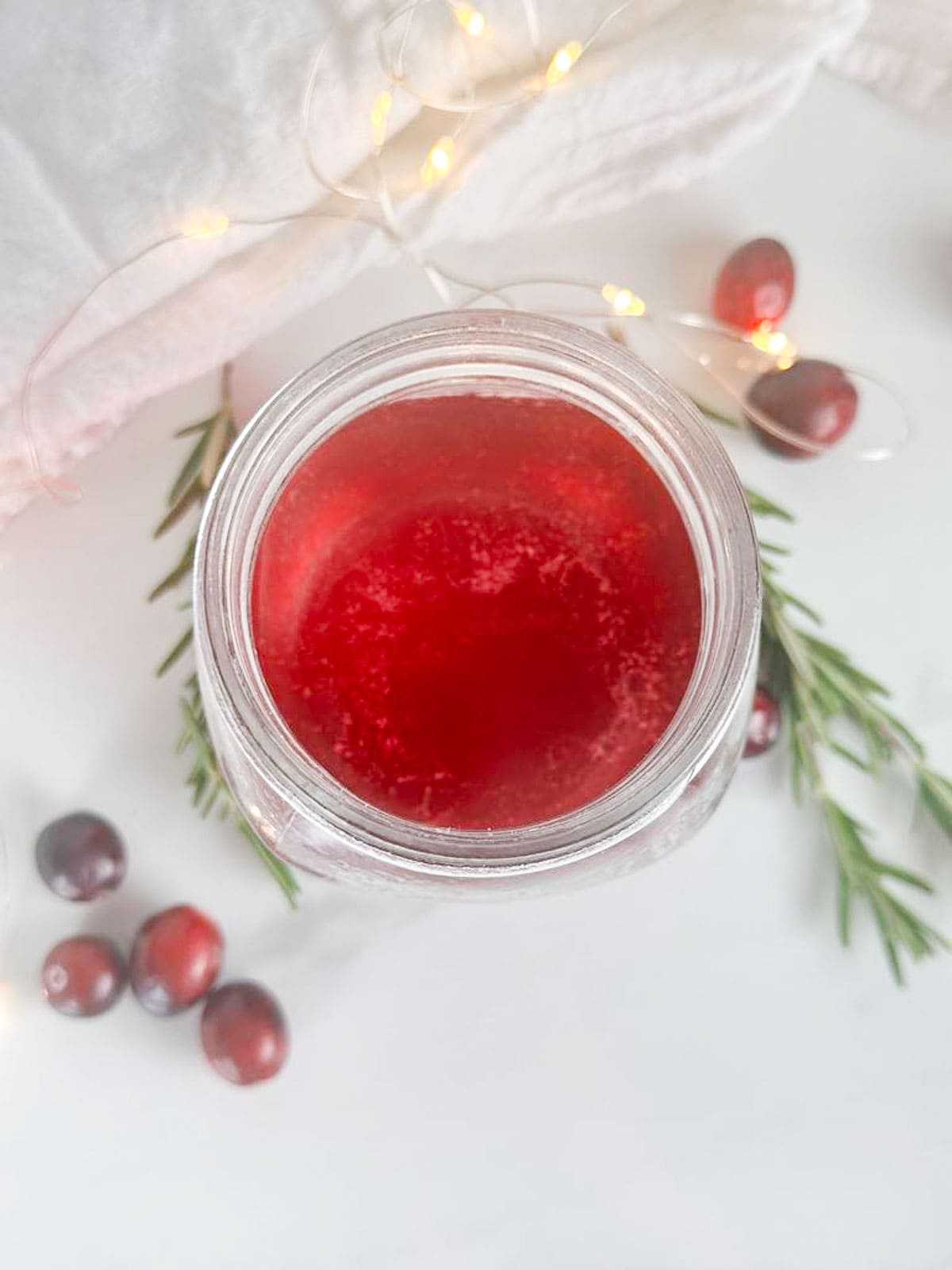 Mason jar without lid with cranberry simple syrup. Cranberries, rosemary sprigs, a tea towel, and twinkle lights are all in the background.