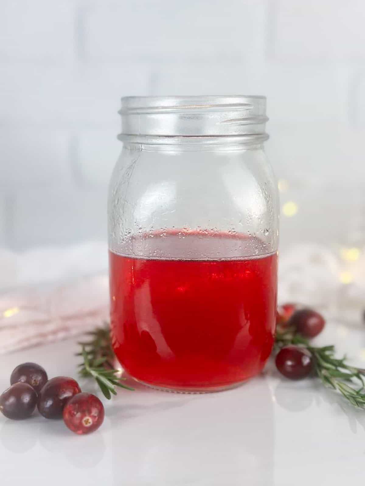 Mason jar without lid with cranberry simple syrup. Cranberries, rosemary sprigs, a tea towel, and twinkle lights are all in the background.