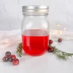 Mason jar with lid with cranberry simple syrup. Cranberries, rosemary sprigs, a tea towel, and twinkle lights are all in the background.