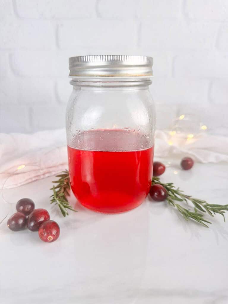 Mason jar with lid with cranberry simple syrup. Cranberries, rosemary sprigs, a tea towel, and twinkle lights are all in the background.