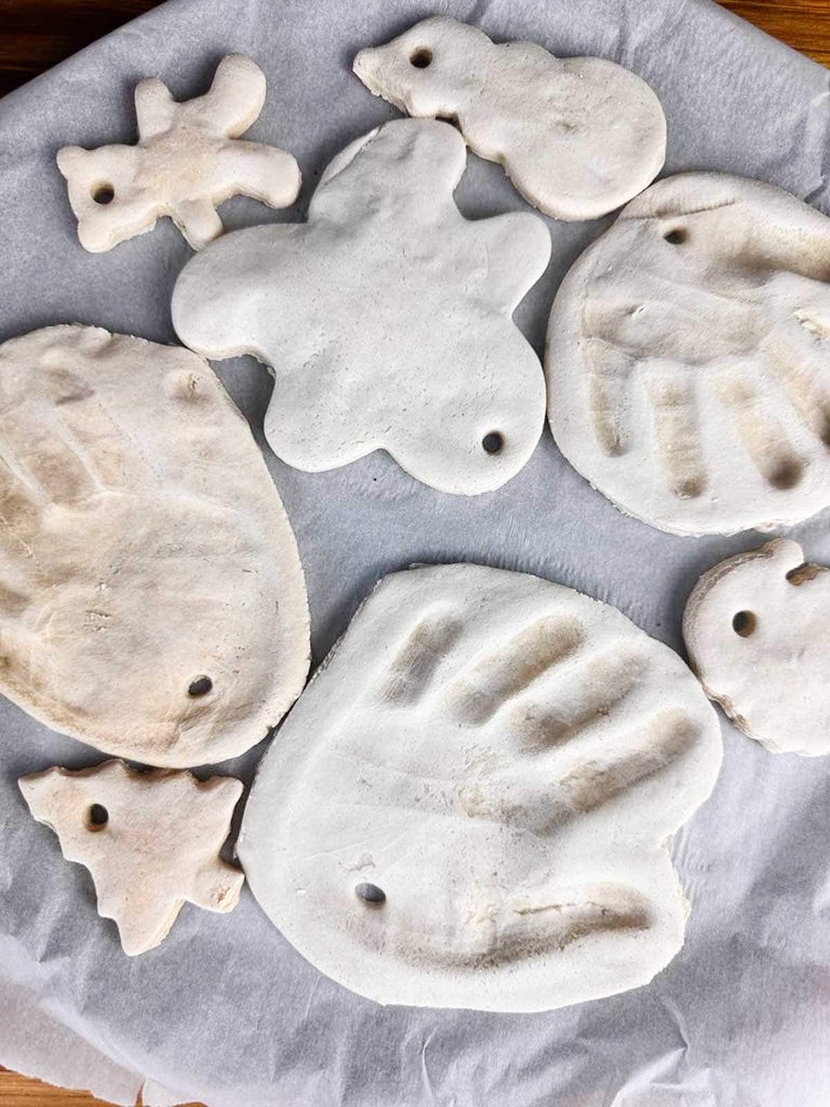 Baked salt dough Christmas ornaments, including: a teddy bear, snowman, gingerbread man, three handprints, Santa, and Christmas tree.