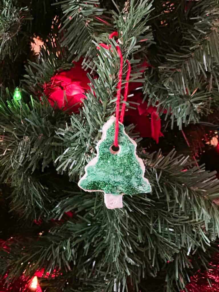 Salt dough Christmas ornament in the shape of a Christmas tree with glitter and red yarn, hanging from a decorated Christmas tree.