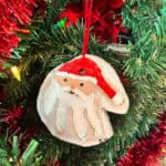 Salt dough Christmas ornament in the shape of a Santa head with red yarn, hanging from a decorated Christmas tree.