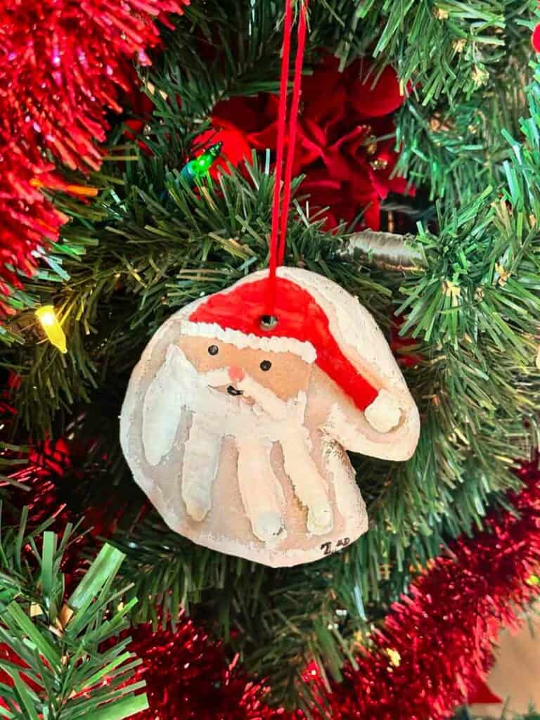 Salt dough Christmas ornament in the shape of a Santa head with red yarn, hanging from a decorated Christmas tree.