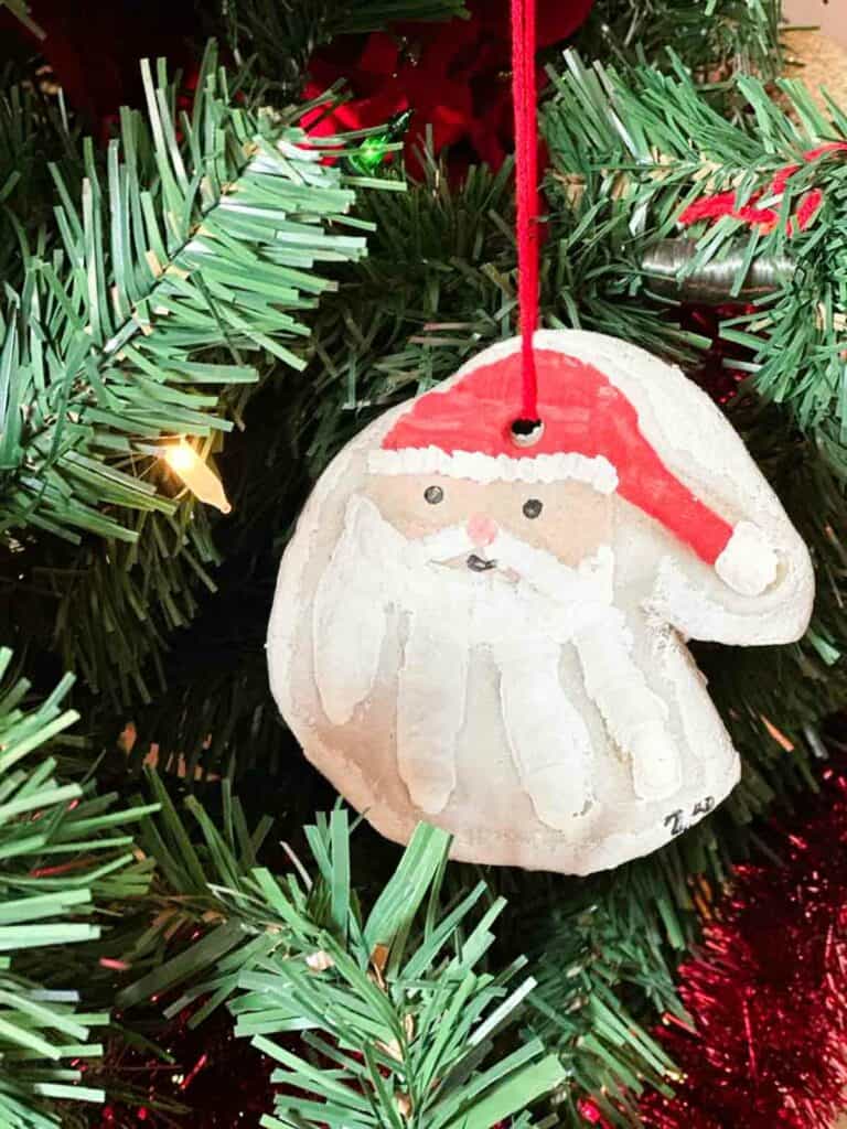 Salt dough Christmas ornament in the shape of a Santa head with red yarn, hanging from a decorated Christmas tree.