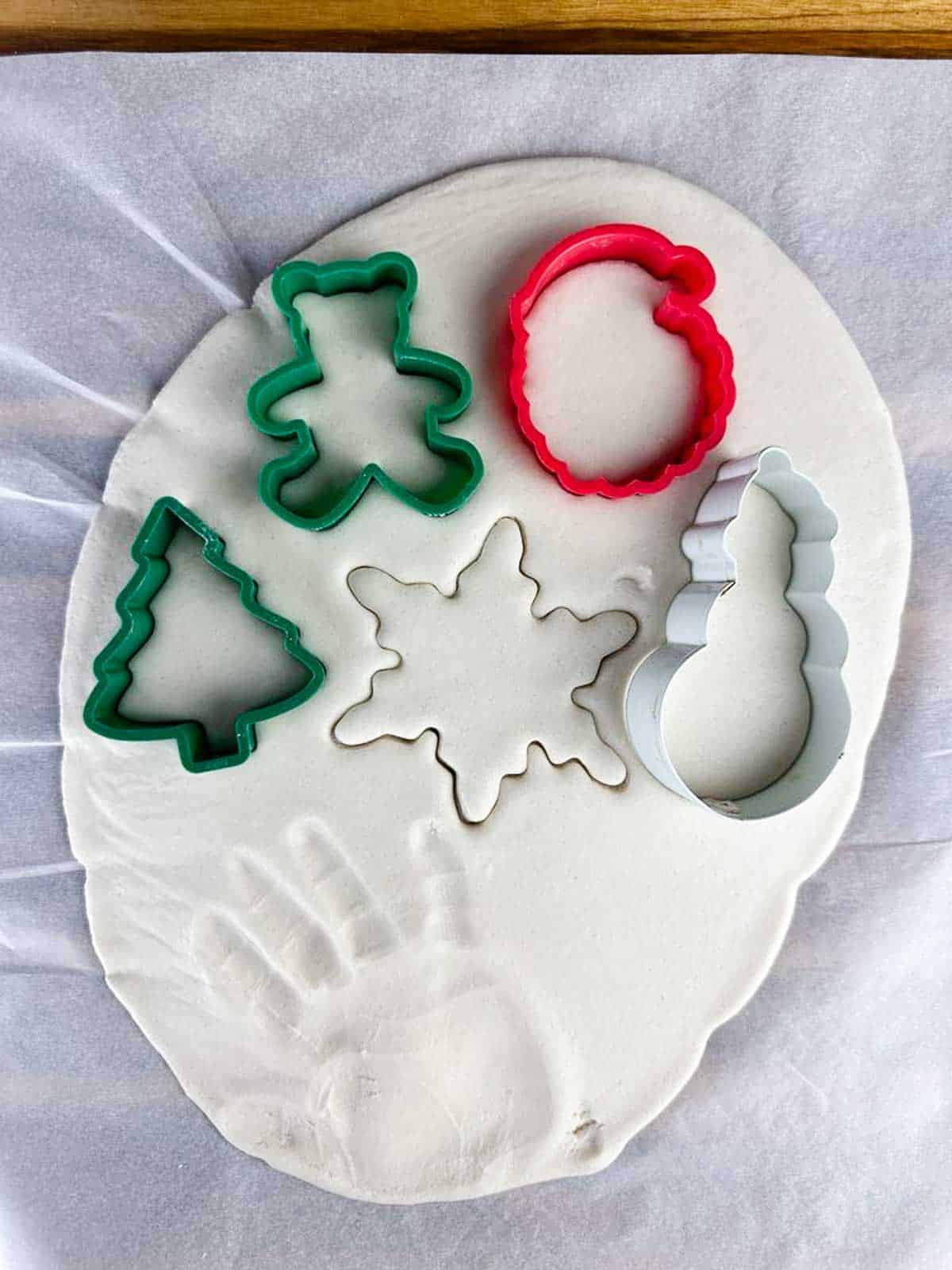 Salt dough for ornaments fully rolled out with a handprint, cut out snowflake, and Christmas tree, teddy bear, santa, and snowman cookie cutters in the dough.