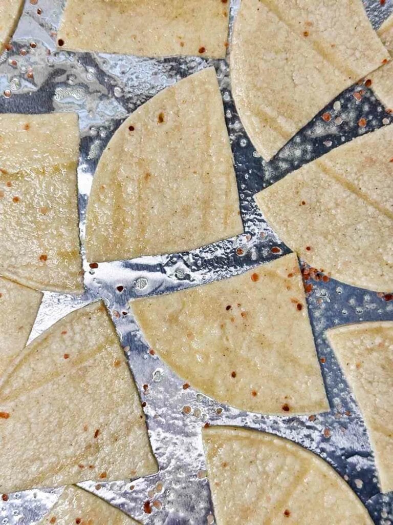 Uncooked corn tortilla chips spread on baking sheet with cooking spray and coarse salt on top.