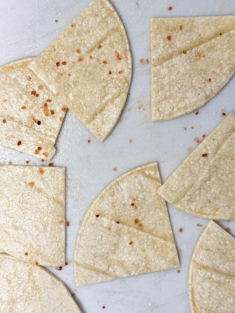 Uncooked corn tortilla chips spread on counter with cooking spray and coarse salt on top, ready for the air fryer.