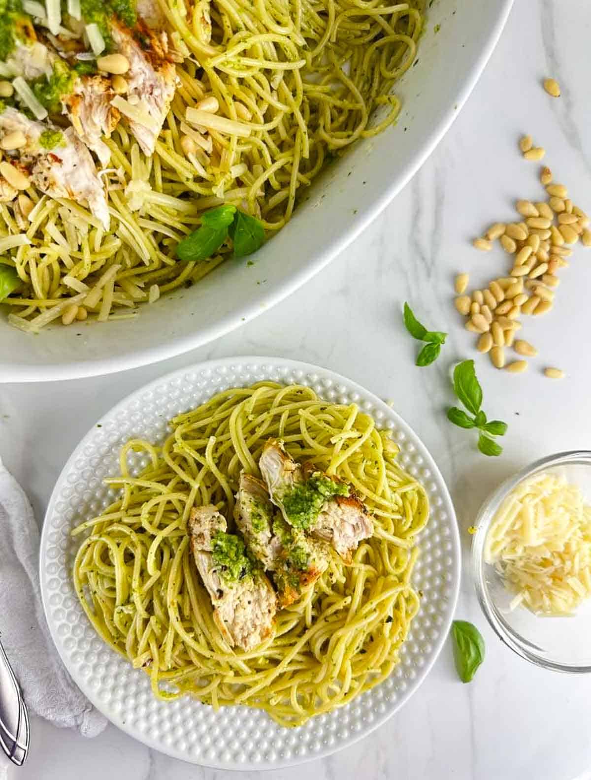 A plate of chicken spaghetti pesto next to a serving platter full of chicken pesto pasta