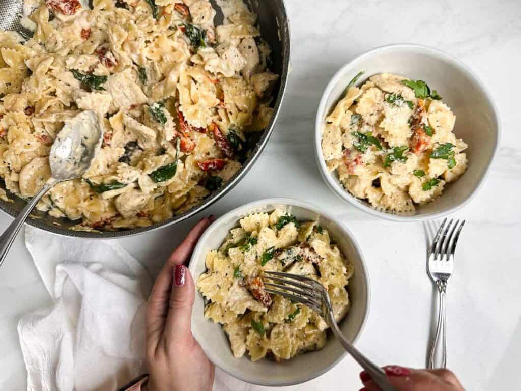 hands holding a fork and bowl to take a forkful of marry me chicken pasta on a set table