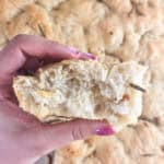 Hand holding a piece of focaccia bread over the rest of the bread.