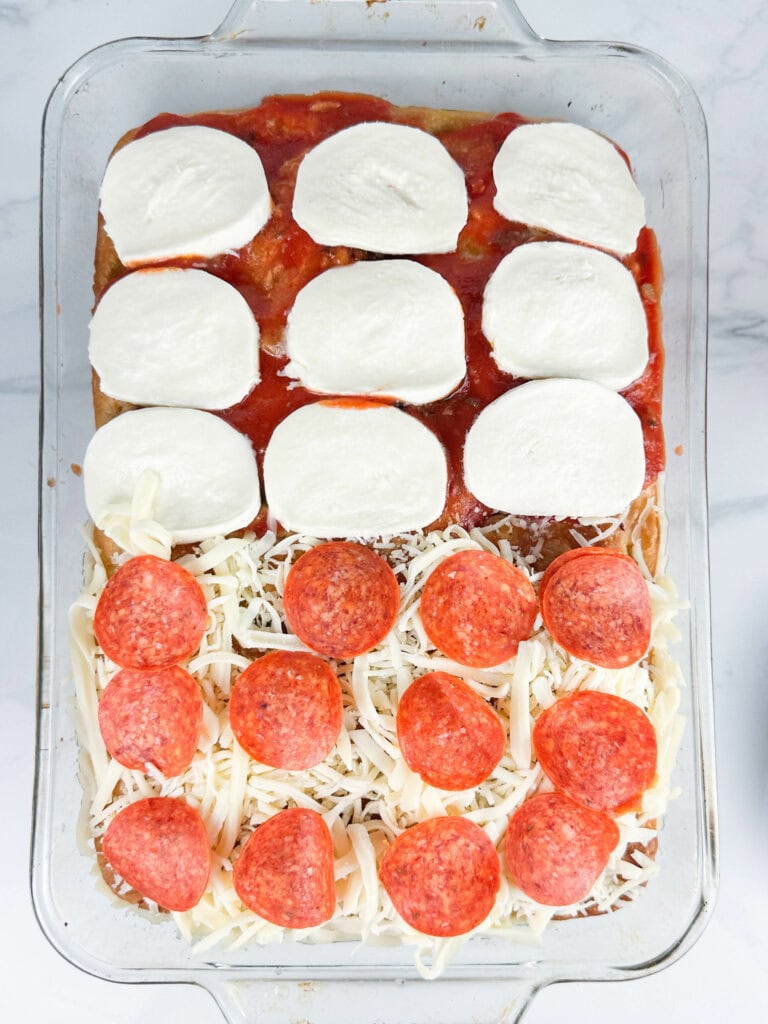 Assembled focaccia bread pizza recipe showing both fresh and shredded mozzarella cheese with pepperoni on top all on sauce set on a neutral background.