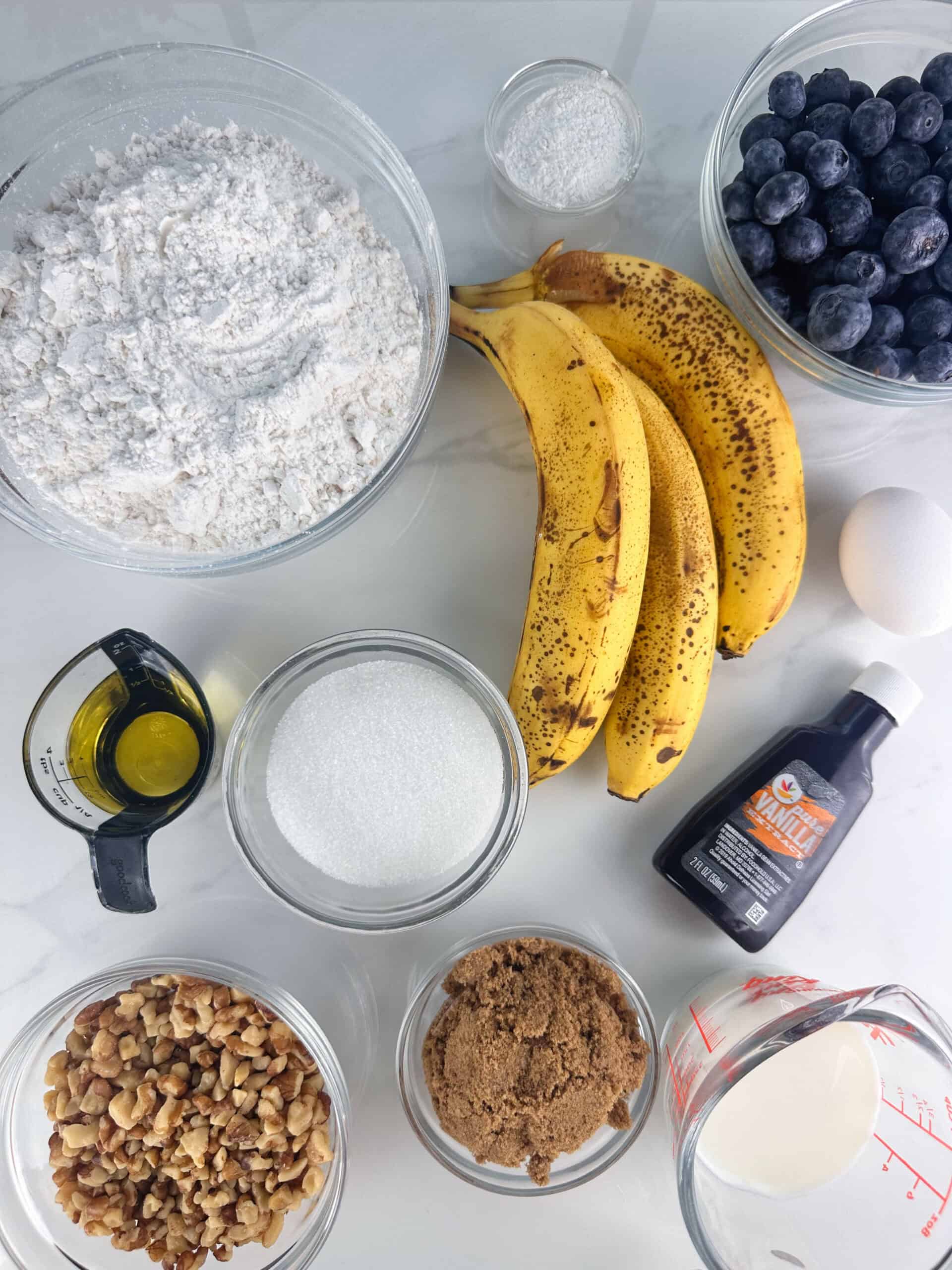 Ingredients for blueberry banana nut bread: flour, baking powder, salt, bananas, blueberries, oil, granulated sugar, vanilla, brown sugar, milk, egg, and walnuts