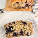 A slice of blueberry banana nut bread on a white plate with the loaf on a wood cutting board