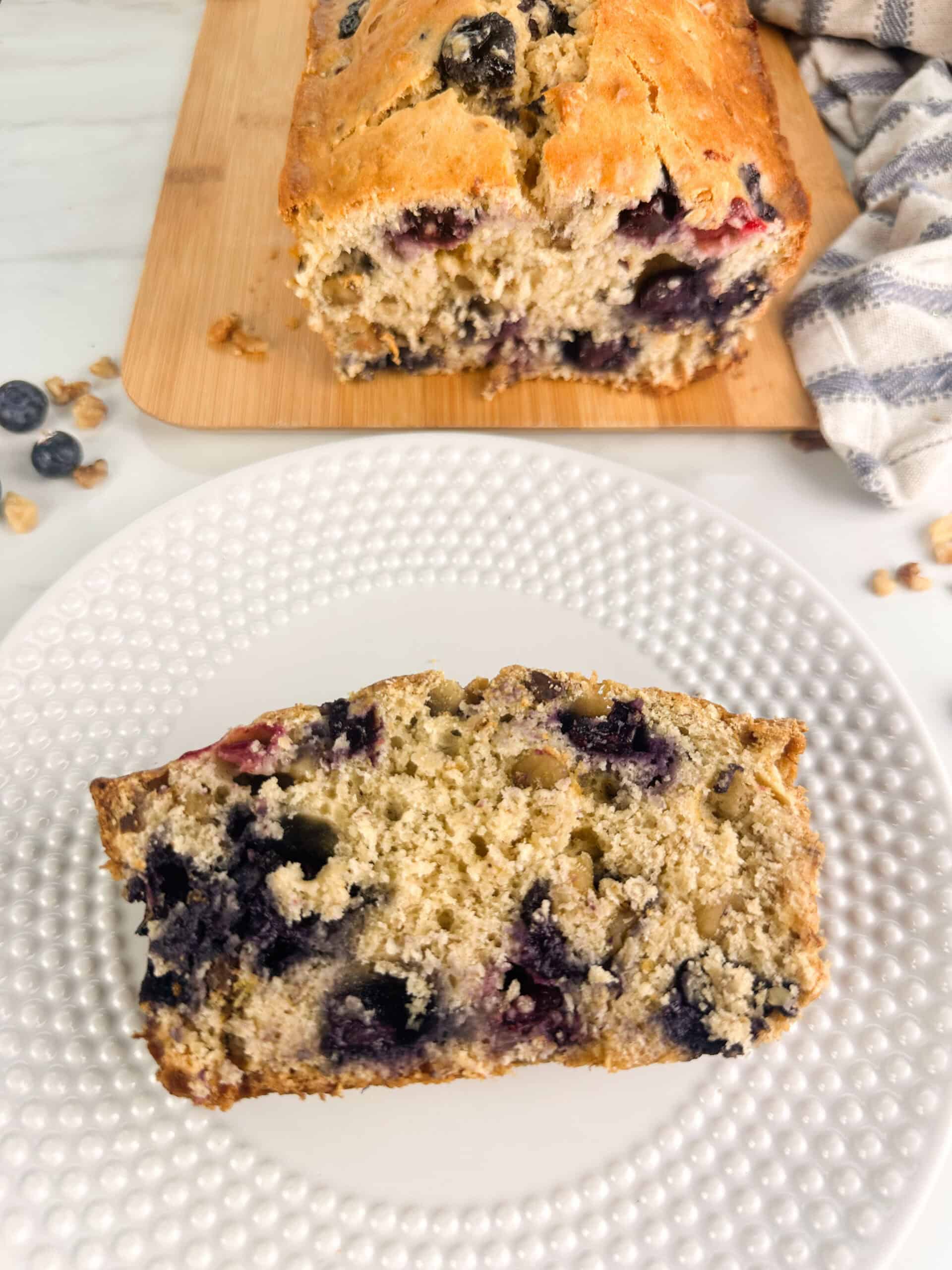 A slice of blueberry banana nut bread on a white plate with the loaf on a wood cutting board