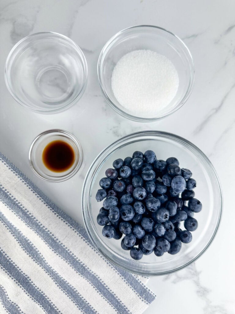 Ingredients for blueberry compote in glass bowls on a white background, ingredients include granulated sugar, blueberries, vanilla extract, and water.