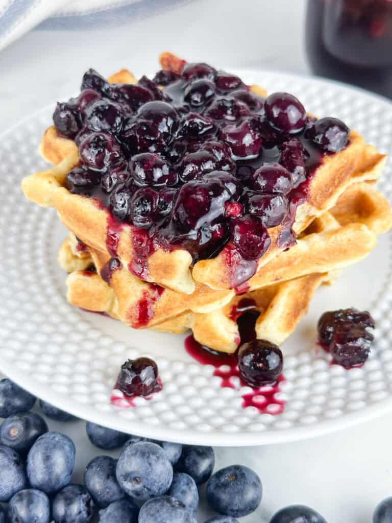 Blueberry compote used as at topping for waffles with blueberries in the front and a jar of the compote in the back on a neutral backdrop.