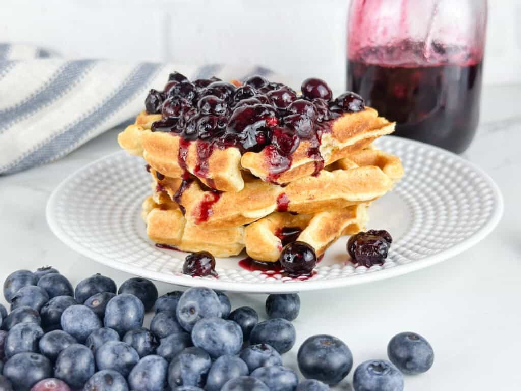 Blueberry compote used as at topping for waffles with blueberries in the front and a jar of the compote in the back on a neutral backdrop.