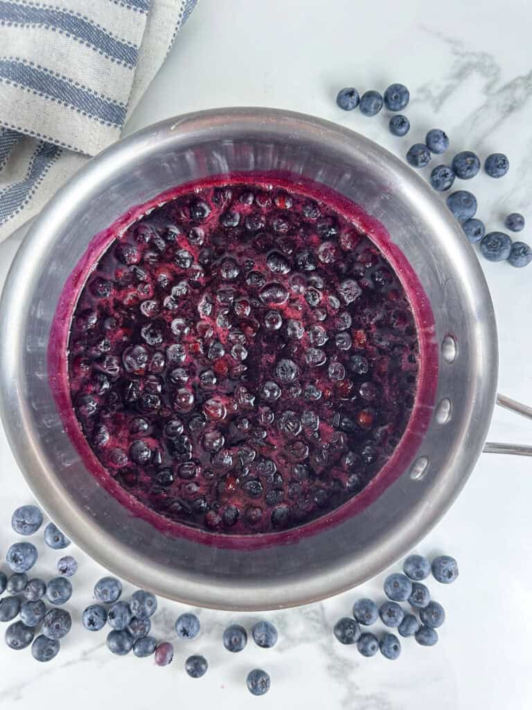 Pot of blueberry compote on a white background with blueberries around the pot.