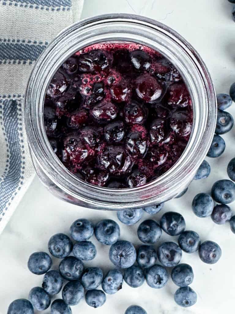 Blueberry compote in a jar on a neutral background with blueberries in the foreground.