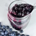 Blueberry compote in a jar on a neutral background with blueberries and spoonful in the foreground.