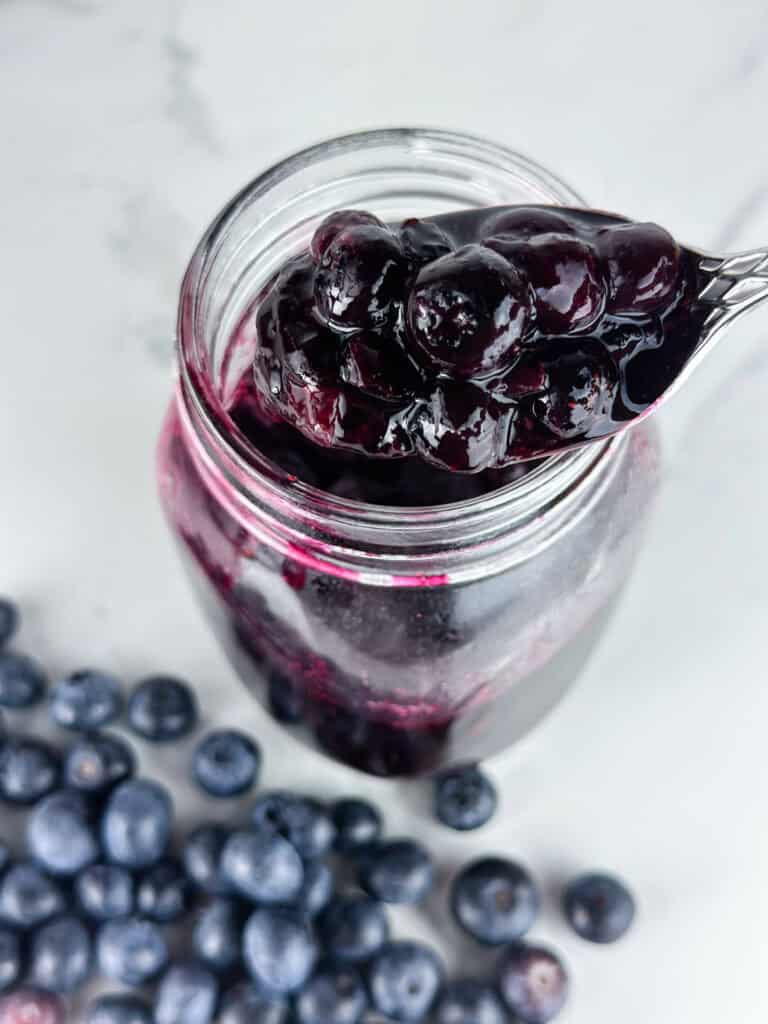 Blueberry compote in a jar on a neutral background with blueberries and spoonful in the foreground.