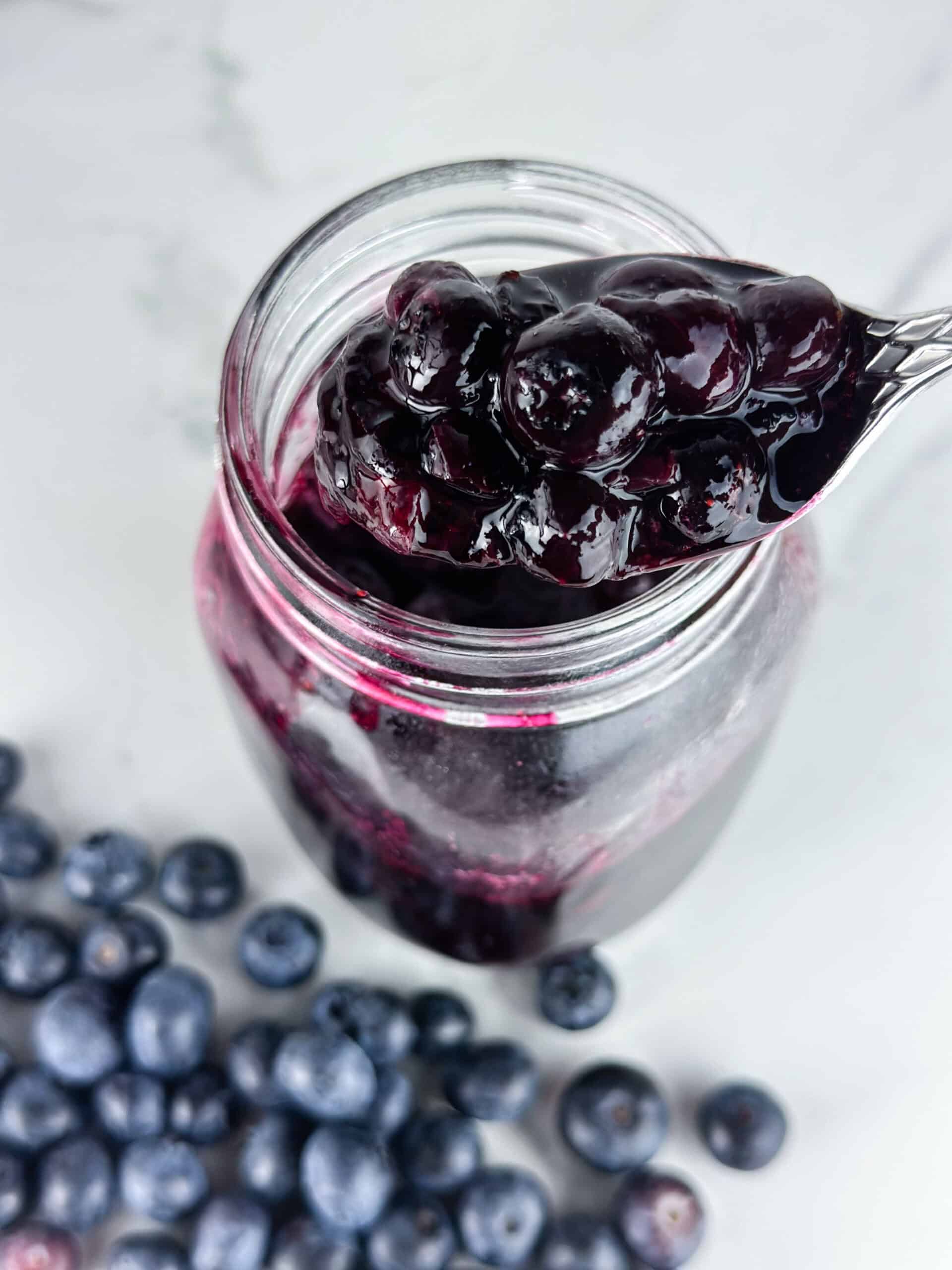 Blueberry compote in a jar on a neutral background with blueberries and spoonful in the foreground.