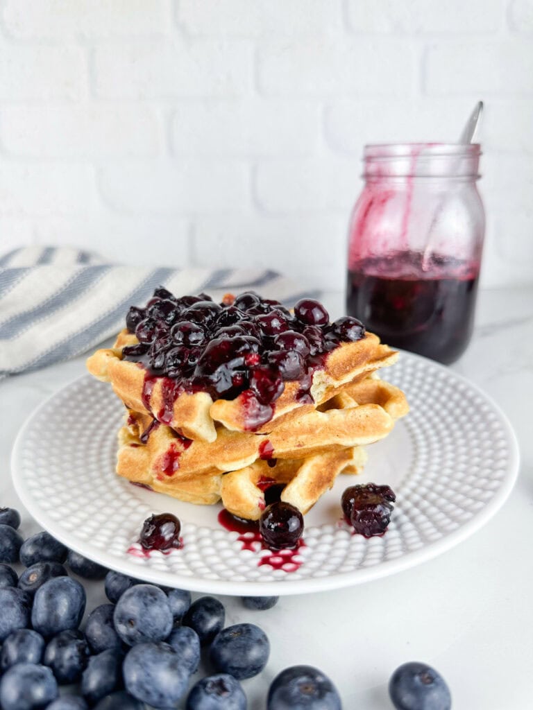 Blueberry compote used as at topping for waffles with blueberries in the front and a jar of the compote in the back on a neutral backdrop.