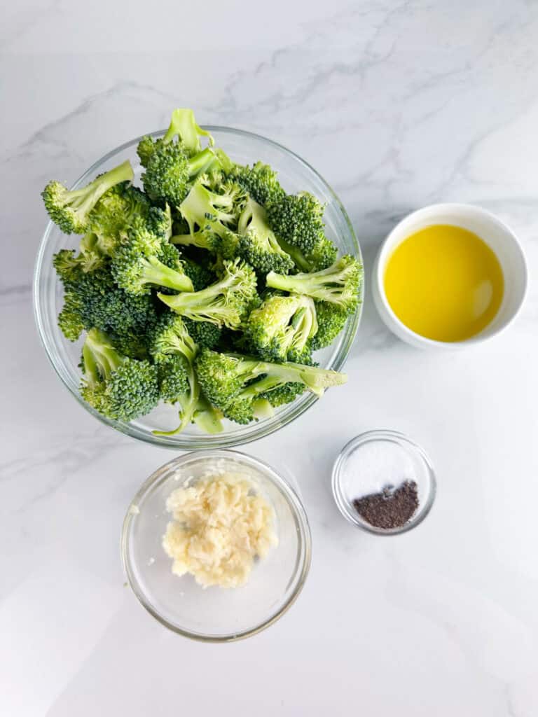 Ingredients for roasted broccoli on a white background, includes: broccoli, olive oil, crushed garlic, salt, and pepper.